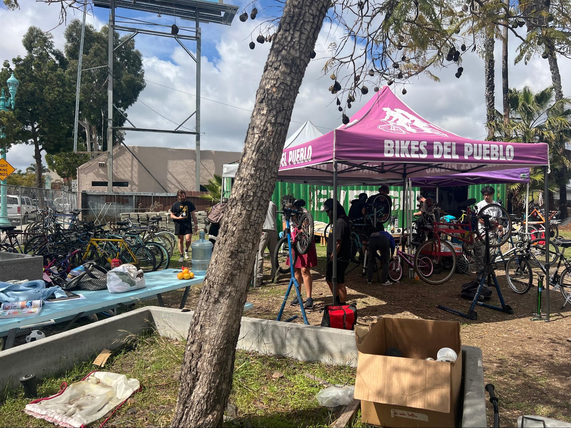 Volunteers and community members mill about the Bikes del Pueblo lot, which borders the I-15 freeway in City Heights. On the left, completed bikes await donation, while volunteers tune up and assemble bikes for community members under the canopy on the right. Photo credit to Dane Persky.